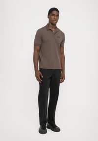 Brun kortermet polo skjorte med krage og brodert logo, sammen med svarte bukser og svarte joggesko, stående på en nøytral bakgrunn.