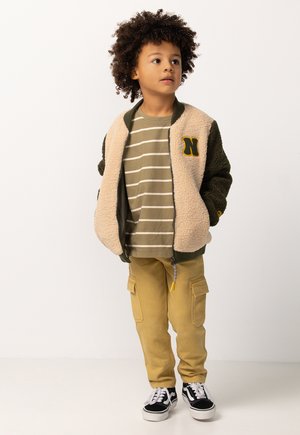 Junges Kind mit lockigem Haar steht, trägt eine beigen und grünen Fleecejacke, gestreiftes Shirt, beige Cargo-Hosen und schwarz-weiße Turnschuhe.