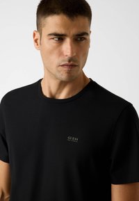 Schwarzes T-Shirt mit kurzen Ärmeln aus geripptem Stoff. Verfügt über einen Rundhalsausschnitt und ein kleines weißes "GUESS"-Logo auf der Brust.