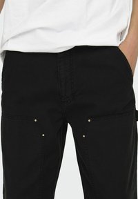 Zwarte cargobroek met een gladde textuur, schuin geplaatste zakken met metalen accenten en een rechte pasvorm. Gestyled met een wit shirt erboven.