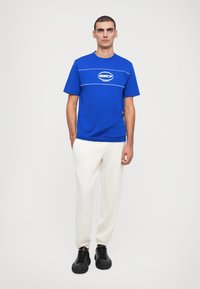 T-shirt bleu à manches courtes avec des lignes et un logo blancs, associé à un pantalon blanc et des chaussures noires à lacets, se tenant devant un fond neutre.