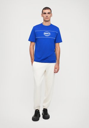 T-shirt bleu à manches courtes avec des lignes et un logo blancs, associé à un pantalon blanc et des chaussures noires à lacets, se tenant devant un fond neutre.