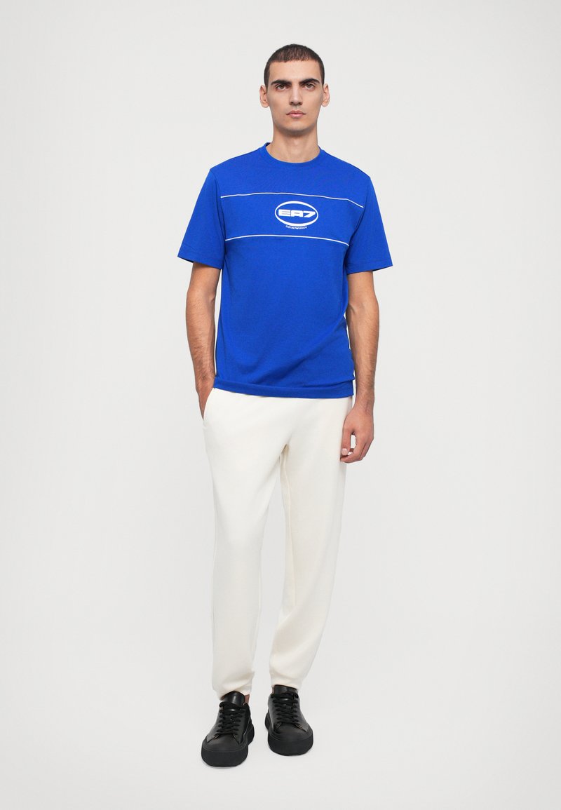 T-shirt bleu à manches courtes avec des lignes et un logo blancs, associé à un pantalon blanc et des chaussures noires à lacets, se tenant devant un fond neutre.