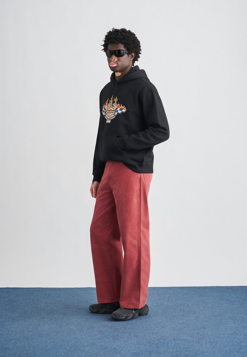 Black hoodie with flame design and logo on chest, paired with wide, red pants and black shoes. Model poses against a neutral background.