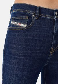 Närbild av en person som bär mörkblå denimjeans med gula sömmar och en liten vit Diesel Industry-etikett på framsidan av fickan.