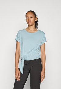Lyseblå t-skjorte med korte ermer og knute i livet. Stoffet er mykt, med en løs passform og rund halslinning, utført sammen med svarte bukser.