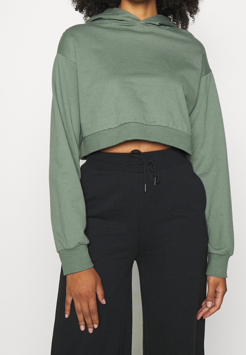 Hoodie vert court avec manches longues et poche frontale, en tissu doux. Associé à un pantalon noir ample avec cordon de serrage.