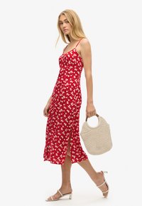 Rotes Midikleid mit Blumenmuster und Spaghettiträgern, seitlichem Schlitz, kombiniert mit einer strukturierten gewebten Tasche und weißen Sandalen mit offenen Zehen.