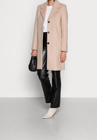 Personne portant un manteau beige à boutons, un haut blanc, un pantalon en cuir noir, des bottes blanches à talons pointus, tenant un sac à main matelassé noir.