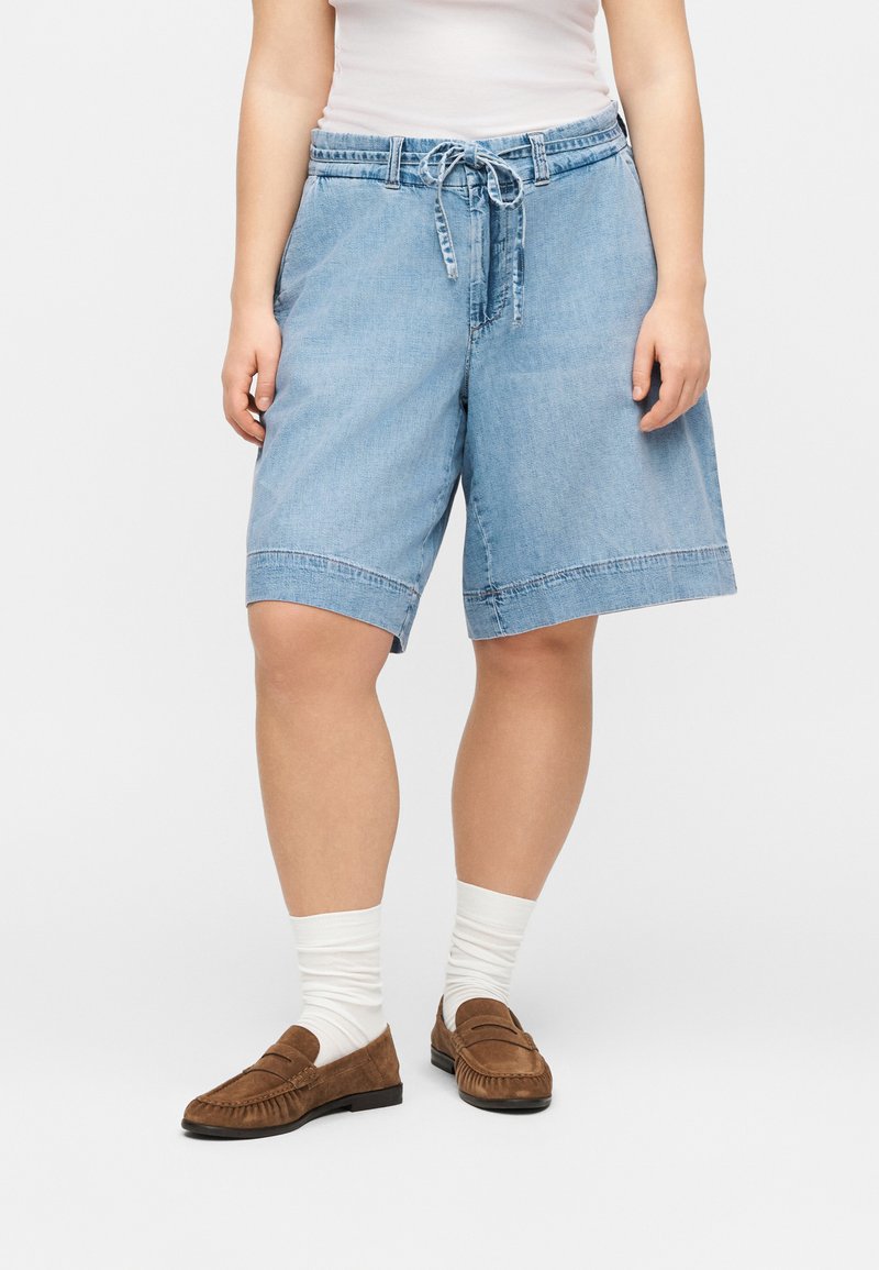 Άτομο που φοράει ανοιχτό μπλε τζιν σορτς με κορδόνι στη μέση, λευκές κάλτσες και καφέ σουέντ μοκασίνια, στέκεται μπροστά σε απλό φόντο.