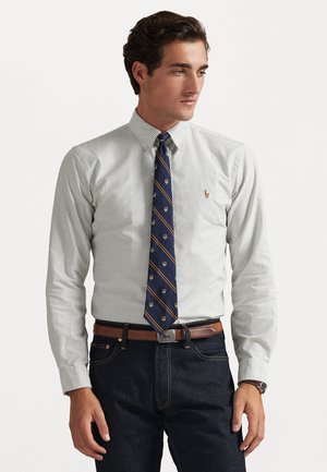 Button-up striped shirt in white and light gray, paired with a navy tie featuring patterns and a brown belt, worn with dark jeans.