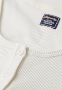 Hvid ribbet tanktop med rund halsudskæring, knaplukning med transparente knapper og et marineblåt label syet på siden.