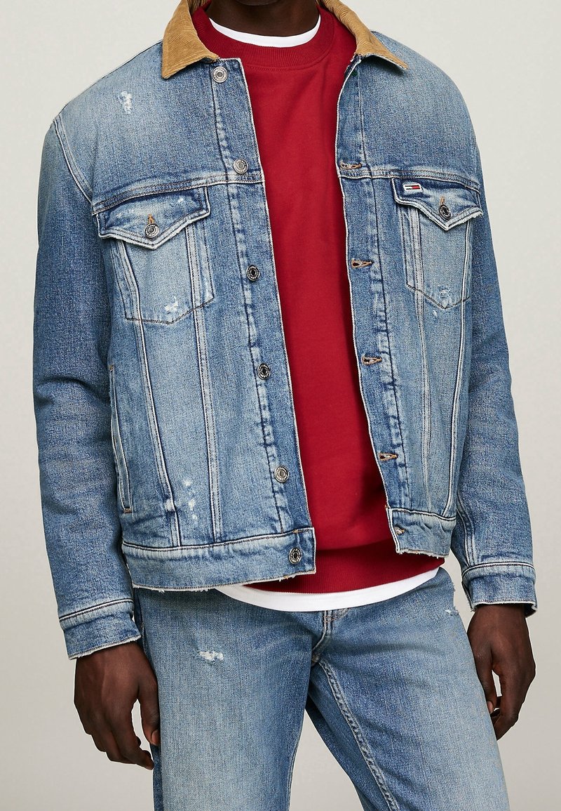 Ljusblå denimjacka med brunt manchesteryta, slitna detaljer, dubbla bröstfickor och knappar. Bärs över en röd sweatshirt.