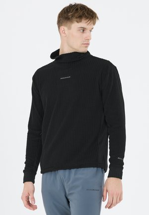 Mand iført en sort tekstureret langærmet top og grå bukser, begge med "Endurance"-logo, kigger til siden mod en ensfarvet baggrund.