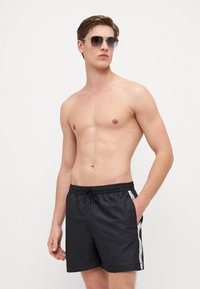 Pantaloni da bagno neri con vita elastica, cordino e strisce laterali che presentano un logo bianco. Modello che indossa occhiali da sole, senza maglietta.