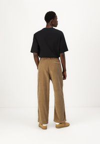 Sort kortærmet t-shirt, brune fløjl vide benbukser med elastisk talje, og beige slip-on sko, set bagfra mod en hvid baggrund.