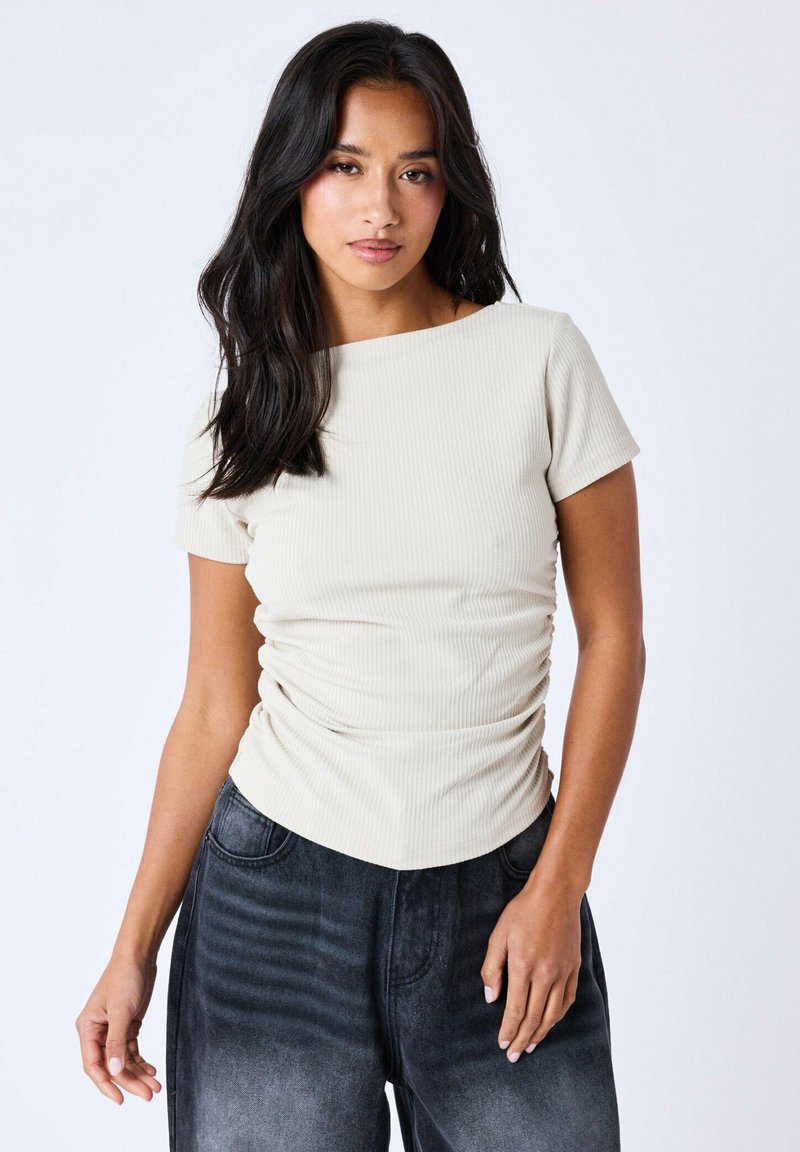 Haut côtelé beige clair à manches courtes, avec des fronces sur les côtés et un décolleté bateau, associé à un pantalon en denim gris foncé.