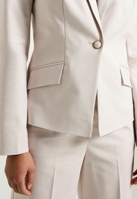 Vaaleanbeige räätälöity puku, jossa on istuva bleiseri yksinkertaisella pyöreällä napilla ja hillityillä taskulapuilla, yhdistettynä samanlaisiin housuihin.