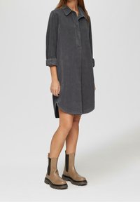 Abito camicia in denim grigio con colletto, maniche lunghe arrotolate, spacchi laterali e vestibilità ampia, abbinato a stivali alla caviglia marroni.