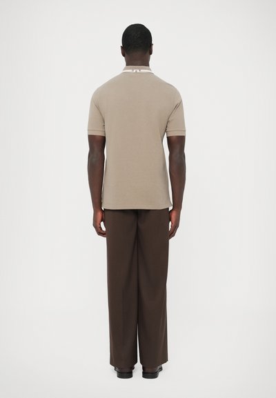 Καφέ κοντομάνικη πόλο μπλούζα με άσπρο γιακά, σε συνδυασμό με καφέ παντελόνια φαρδιάς γραμμής. Το μοντέλο στέκεται με την πλάτη γυρισμένη προς φωτεινό φόντο.