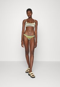 Biquíni de dois pedaços riscado em verde oliva e branco, apresentando alças finas e um design mínimo, combinado com sandálias de slide de cor clara com babados.
