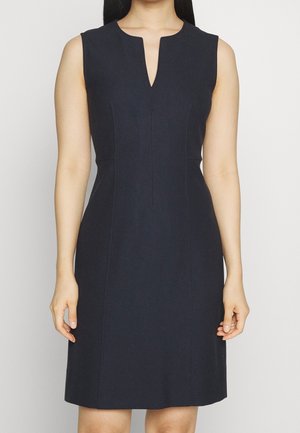 Hihaton laivastonsininen mekko, jossa on istuva vyötärö ja edessä V-mallinen kaula-aukko, pukeutuneena henkilö, joka seisoo rento asento käsivarret alhaalla.