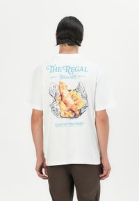 White short-sleeve t-shirt featuring graphic print of fish and chips on the back, with blue text reading "The Regal" and "Let’s Get Battered."