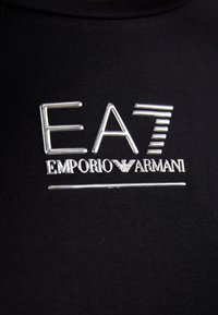 Zwarte stof met een gladde textuur heeft een zilveren metallic logo met de tekst "EA7 EMPORIO ARMANI" en horizontale en verticale ontwerpaccenten.