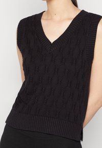 Vrouw die een mouwloze zwarte kabelgebreide V-halstrui zonder mouwen draagt, gecombineerd met zwarte broek, waarbij de bovenste romp en nek zichtbaar zijn.