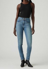 Jeans ajustados en denim azul claro, con una cintura alta, diseño de cinco bolsillos y un ligero desgaste, combinados con una camiseta sin mangas negra y botas negras hasta el tobillo.