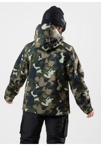 Kamouflagejacka i gröna, bruna och svarta nyanser; har en huva, elastiska muddar och en liten logotyp på baksidan. Bärs med svarta byxor.