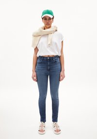 Top corto blanco de manga corta, jeans ajustados azul de cintura alta, suéter de punto blanco roto alrededor del cuello, gorra verde y sandalias blancas con tiras.