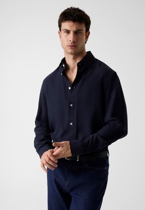 Junger Mann mit lockigem Haar, trägt ein dunkelblaues Hemd mit Knöpfen und dunkle Hosen, steht vor einem schlichten hellen Hintergrund.