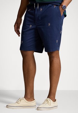 Man draagt marineblauwe korte broek met kleine witte en rode geborduurde motieven en beige vetersneakers tegen een effen achtergrond.