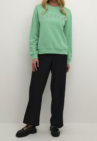 Sudadera verde con el texto "EXHALE" en blanco, combinada con pantalones negros holgados. El calzado presenta una suela negra gruesa y acentos en dorado.