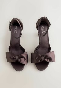 Bruine satijnen hoge hakken sandalen met enkelbandje en een grote strikdetail aan de voorkant. Gladde textuur met een glanzende afwerking.