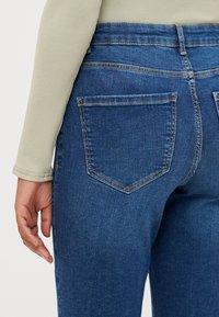 Vaqueros de mezclilla azul oscuro con pierna ajustada, con una cintura de altura media, bolsillos traseros y una textura suave. Combinados con una blusa de color verde claro.