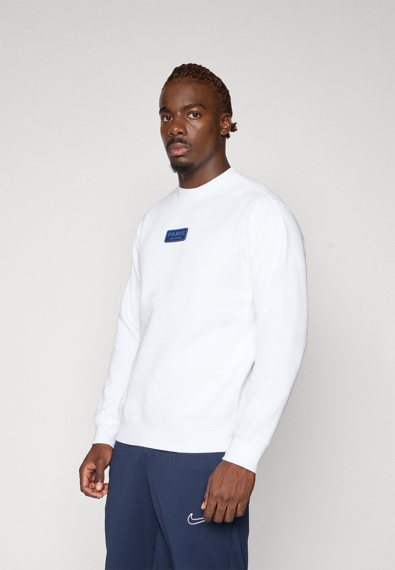 Sweatshirt blanc à col rond avec une pièce rectangulaire bleue sur laquelle est inscrit "PARIS". Le tissu semble lisse avec des poignets et un ourlet côtelés.