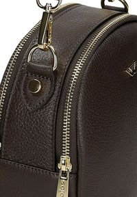 Sezione di borsa a tracolla in pelle marrone scuro texturizzata con cerniera dorata, chiusura in metallo e piccolo logo su una superficie curva.