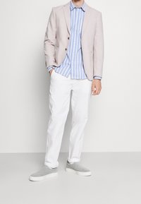 Mężczyzna w beżowej marynarce, niebieskiej i białej koszuli w paski z guzikami, białych spodniach i szarych butach bez pięty stoi na jasnym tle.