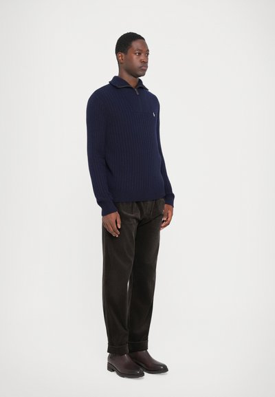 Suéter de punto acanalado en navy con cuello con cremallera, combinado con pantalones de pana oscuros y botas oscuras, creando un look texturado y en capas.