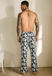 Shirtless man with tattoos on arms and upper back wearing black and white ghost-print pajama pants standing barefoot on wooden floor.