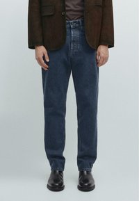 Jeans blu scuri con taglio dritto, in tessuto denim, e cuciture visibili, abbinati a un blazer marrone e stivali in pelle nera.