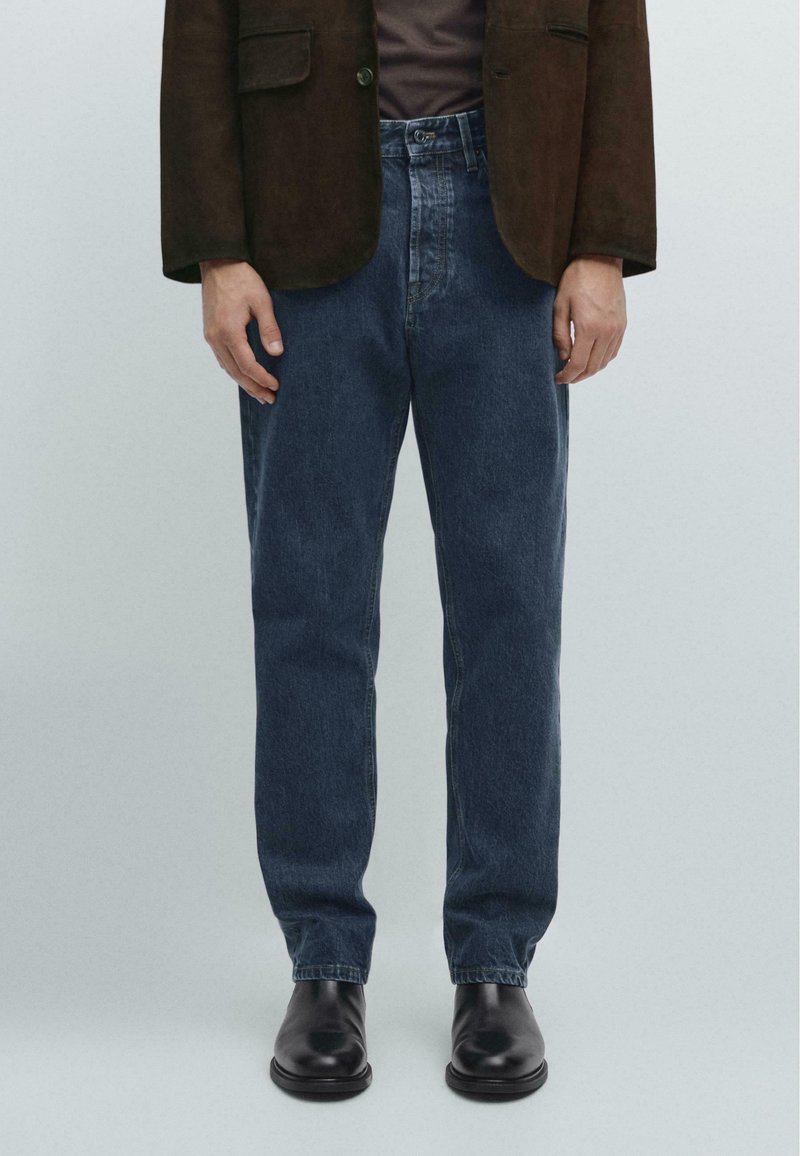 Jeans blu scuri con taglio dritto, in tessuto denim, e cuciture visibili, abbinati a un blazer marrone e stivali in pelle nera.