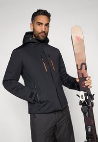 Čierna lyžiarska bunda s oranžovými zipsami a textúrovaným ramenným panelom, v ruke drží lyžu a viazanie; čierne nohavice dokončujú outfit.