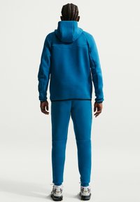 Μπλε φούτερ με κουκούλα και ταιριαστό παντελόνι, κατασκευασμένα από απαλό υλικό. Σημαντικές λεπτομέρειες είναι οι αντίθετες μαύρες ραφές και η εφαρμοστή σχεδίαση.