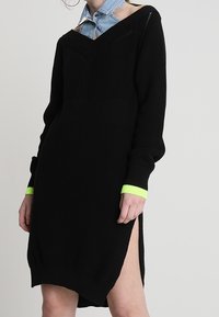 Svart stickad klänning med långa ärmar, V-ringning och sidoklyftor. Har en neongrön accent vid ärmkanterna, texturerad tyg och löst sittande passform.