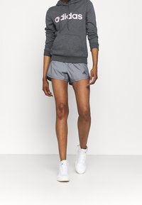 Szara bluza z kapturem z różowym logo "adidas", zestawiona z szarymi szortami sportowymi z czarnymi akcentami i białymi trampkami. Luźny strój sportowy.