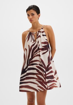 Femme portant une robe courte sans manches, ample, avec des motifs abstraits de feuilles marron et noir sur fond blanc.