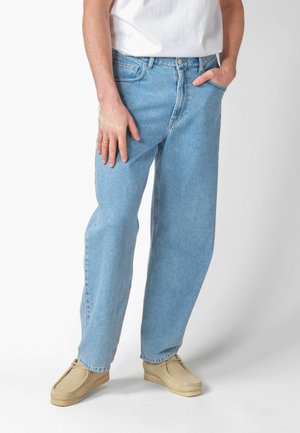 Person iført lyseblå jeans med lige ben, hvid skjorte og beige mokkasinstil sko, står med den ene hånd i lommen og den anden hvilende på låret.